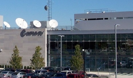 Broadcast headquarters featuring multiple satellite dishes atop a modern building, surrounded by a parking lot filled with cars.